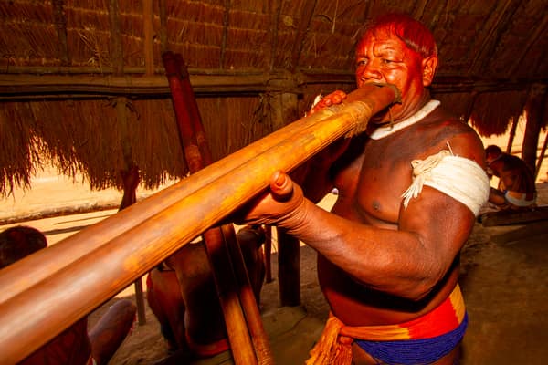 An elder playing a long wooden flute inside a thatched house