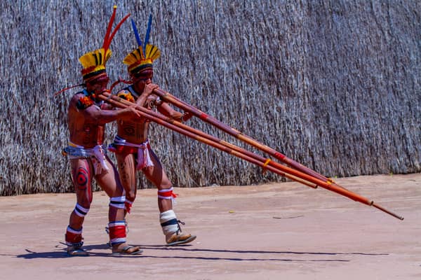 Two men playing long flutes before a large thatched house